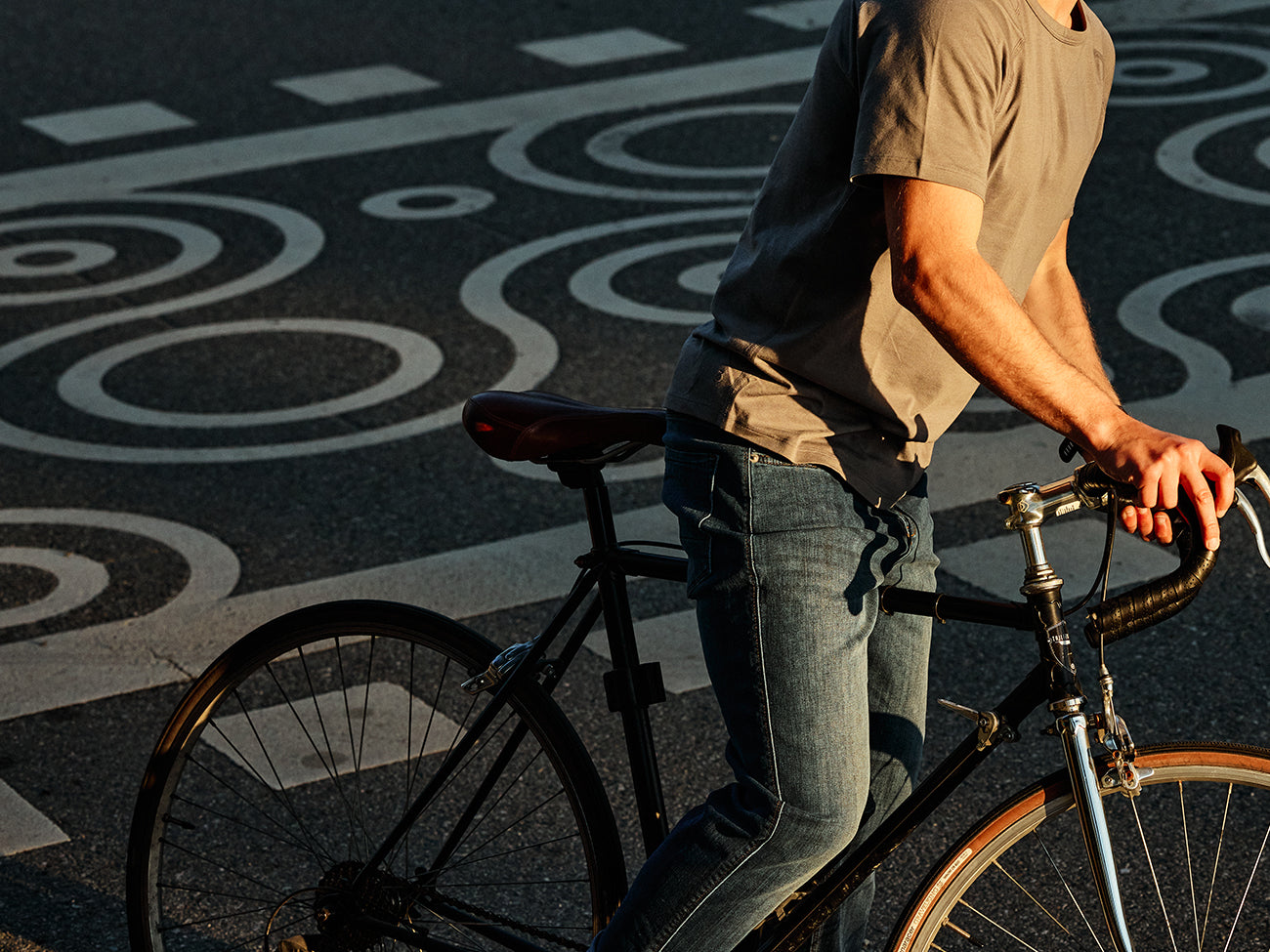 A person wearing a grey t-shirt and blue jeans is riding a bicycle on a street with artistic patterns.