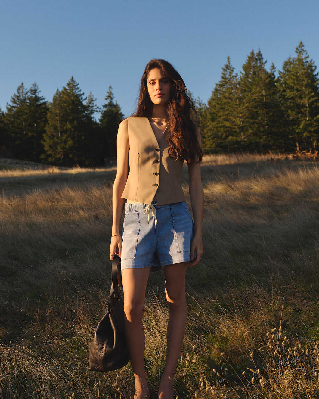 A woman stands outdoors in a grassy field. Wearing a beige sleeveless buttoned top and denim shorts, holds a black bag in her hand.