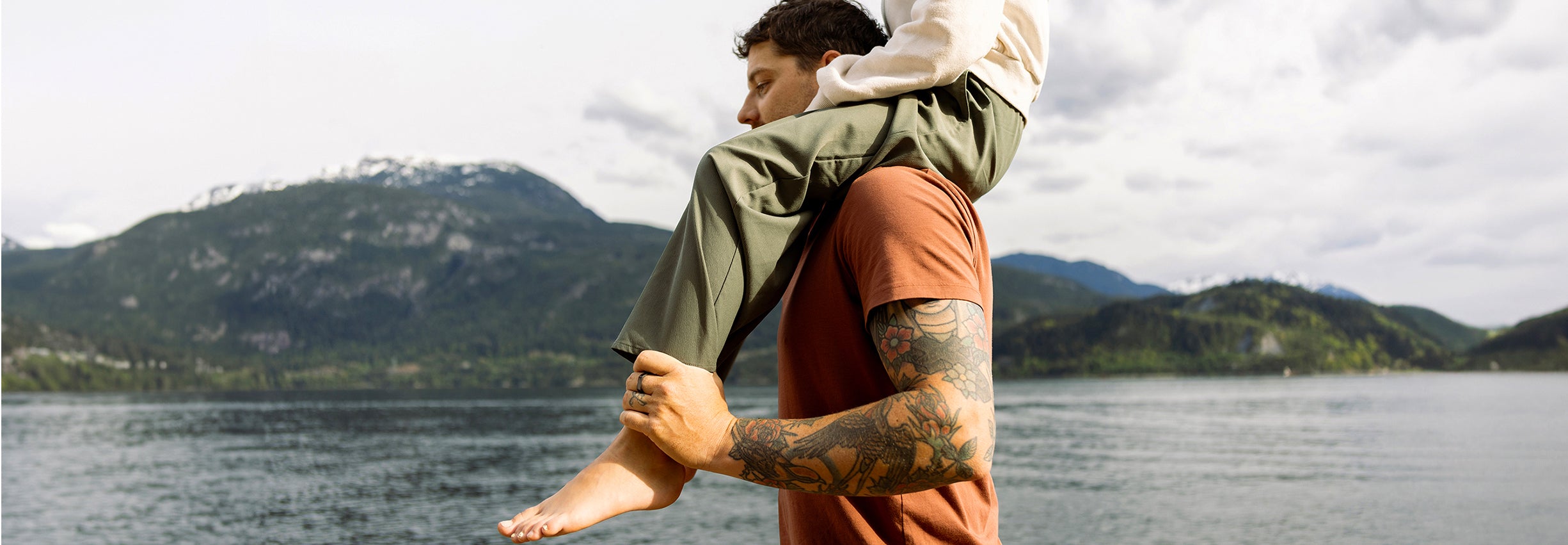 A man with arm tattoos carries a woman on his shoulders by a large scenic lake.