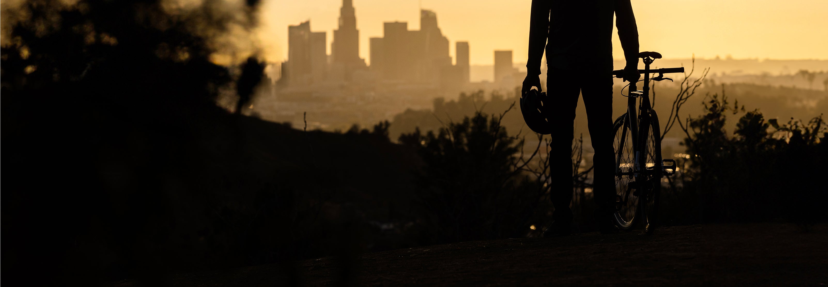 Silhouette of man holding a bike helmet and standing beside a bike while over looking a city skyline