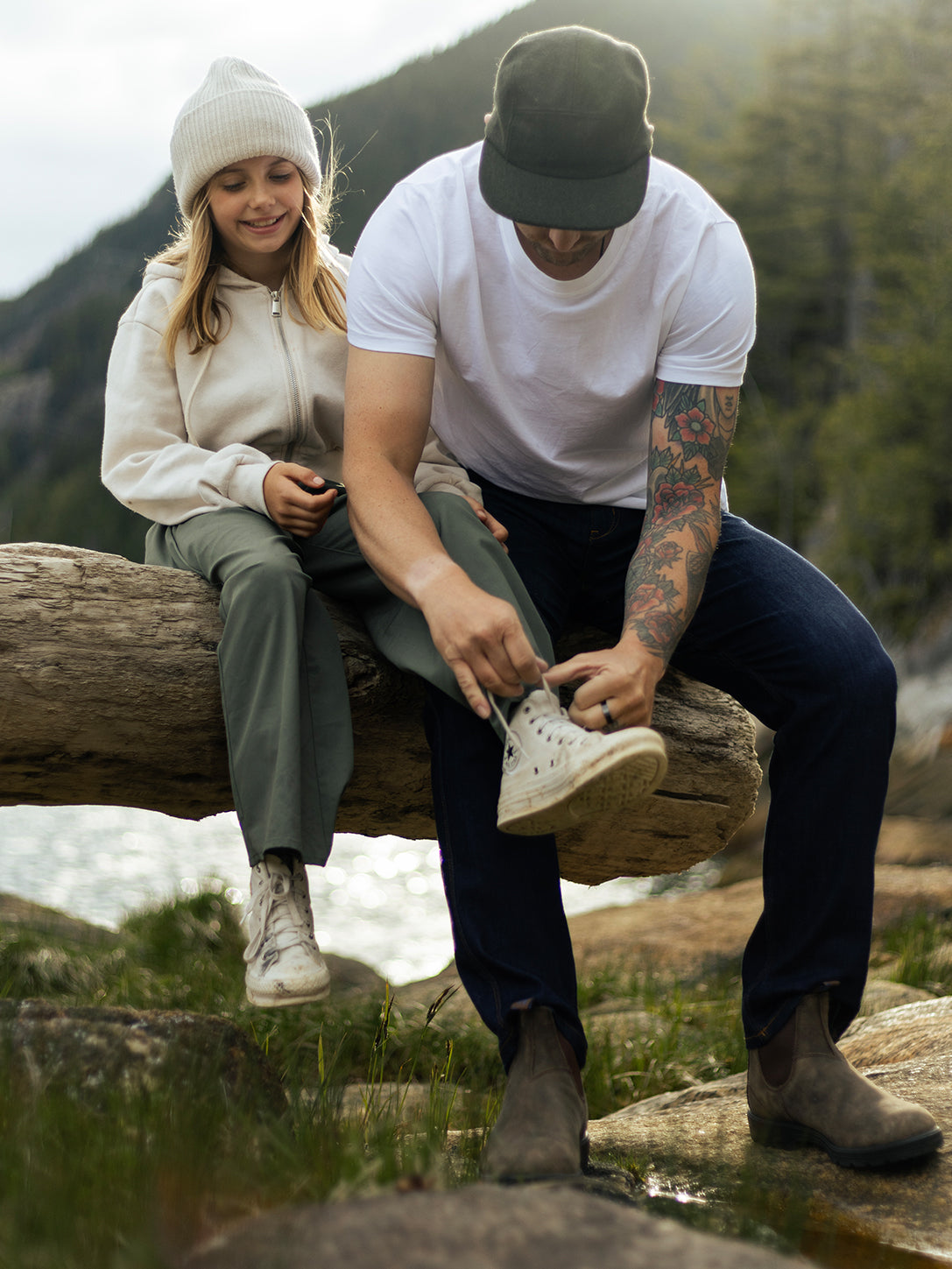 Man tying shoelaces of smiling girl on a log near water, with trees and hills in the background