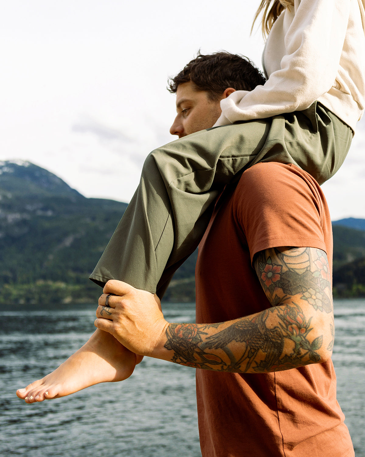 A man with arm tattoos carries a woman on his shoulders by a large scenic lake.