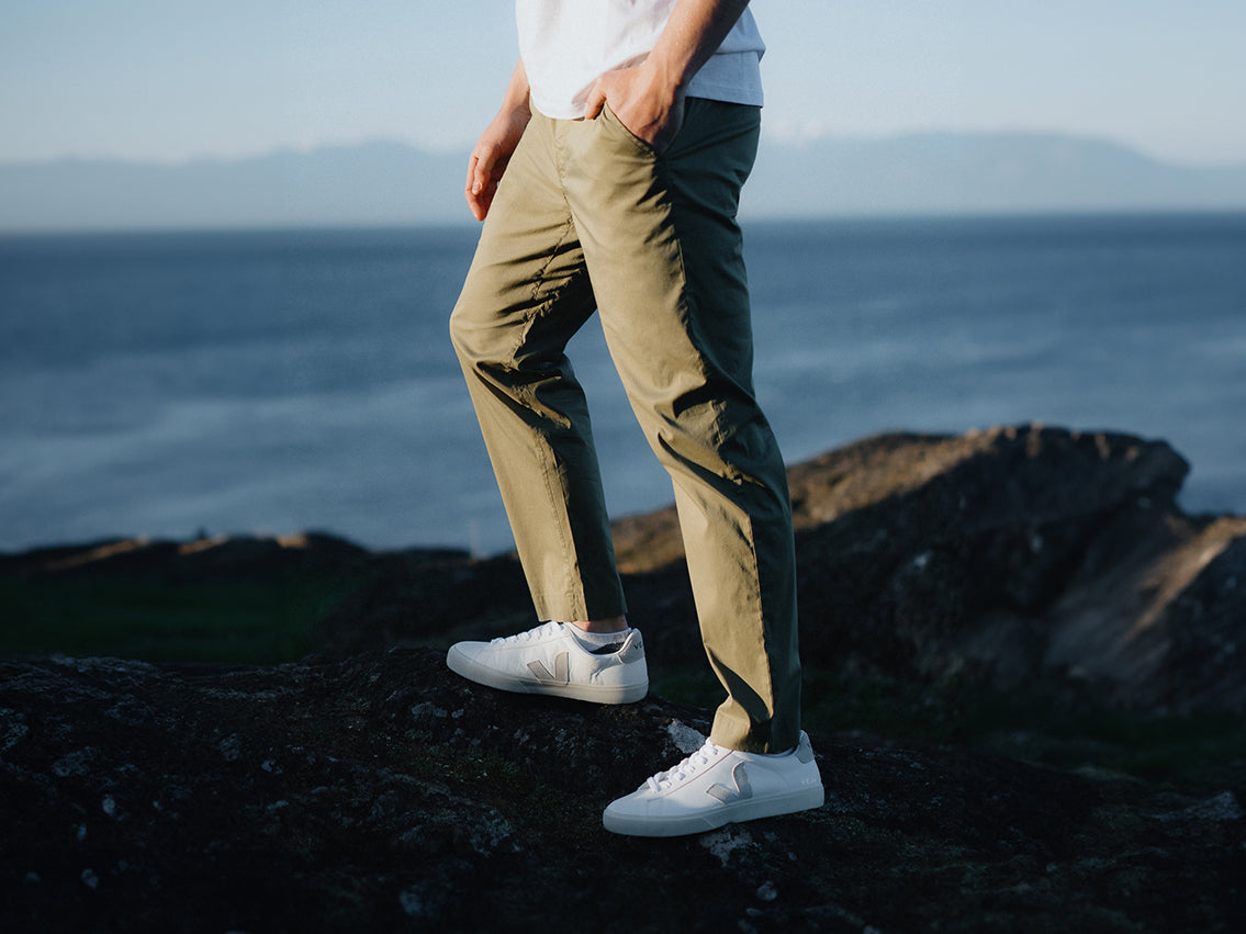 A man wearing green pants and white shirt stands on rocks near body of water. 