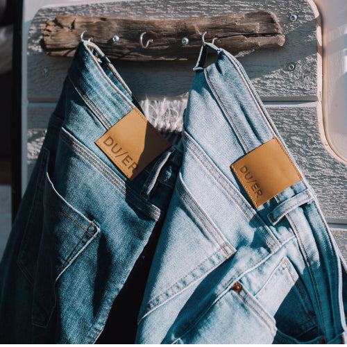 two pairs of DUER jeans hanging by their belt loops on wood hooks