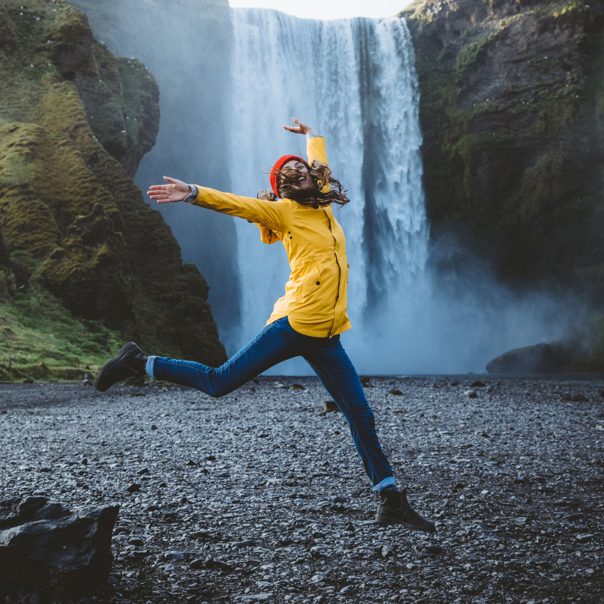 woman jumping in a yellow raincoat in front of a waterfall