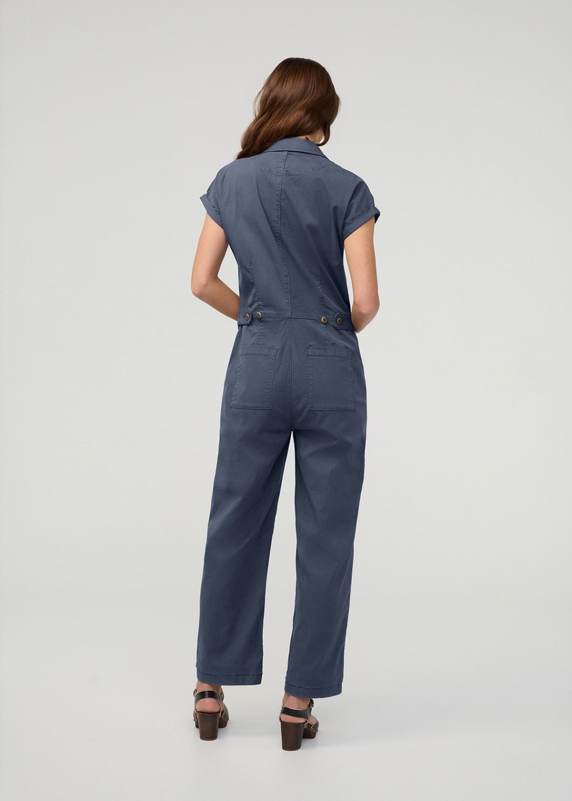 Woman wearing heritage blue short-sleeved boiler suit with front pockets and button closure, shown from behind.