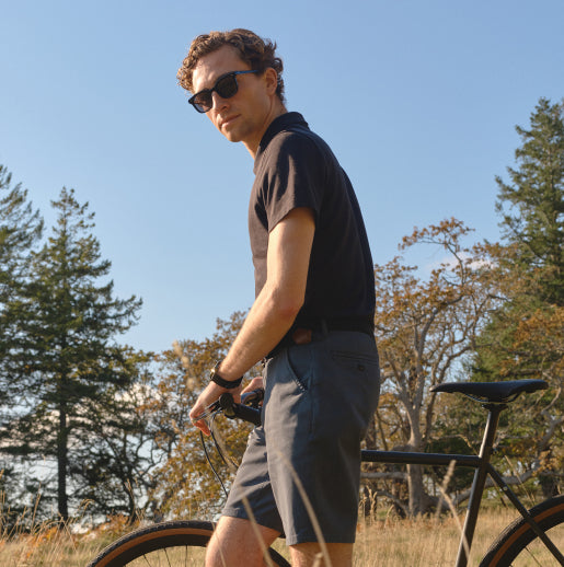 A man wearing a black polo shirt, grey shorts and sunglasses, walking his bike through a field. 