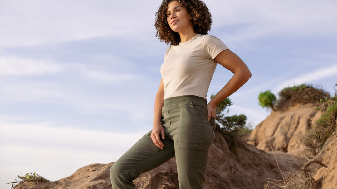 woman standing outdoors against a rocky landscape, wearing a beige shirt and olive green pants.