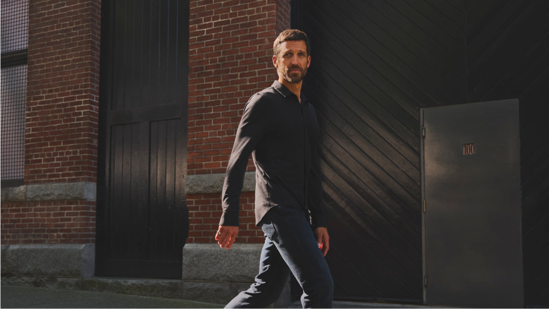 A man dressed in a dark collared shirt and jeans, walking past a brick building.