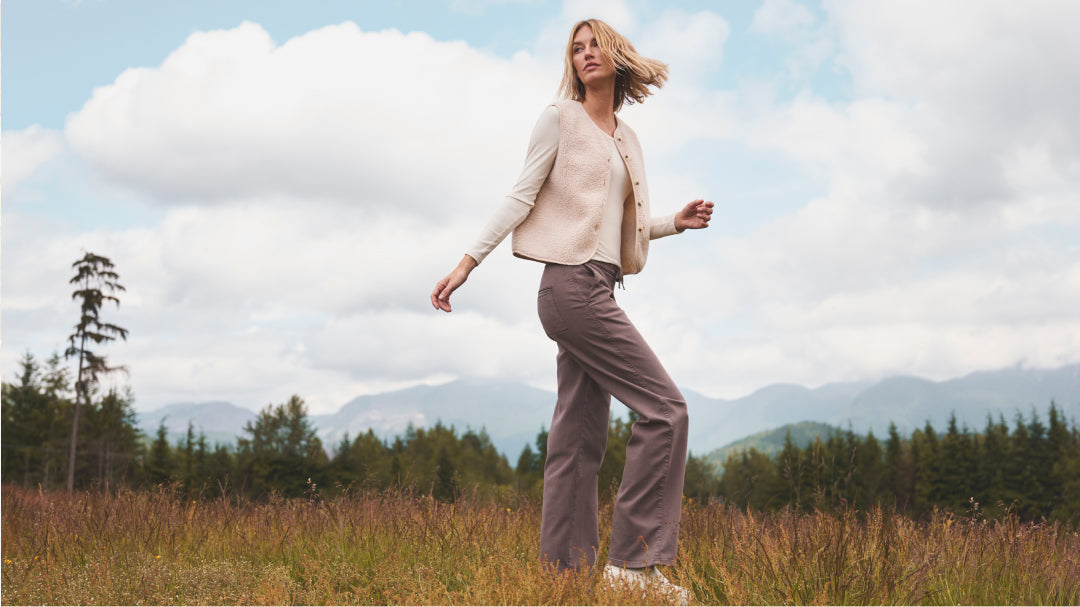 A woman stands in a grassy field, wearing a beige vest, long-sleeved shirt, and brown wide leg pants.