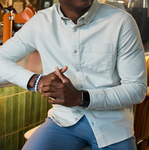 A man sitting on a bar stool, dressed in blue pants and a white collared long-sleeve shirt