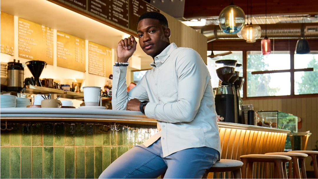 A man seated on a bar stool at a café, wearing blue pants and a white collared long-sleeve shirt