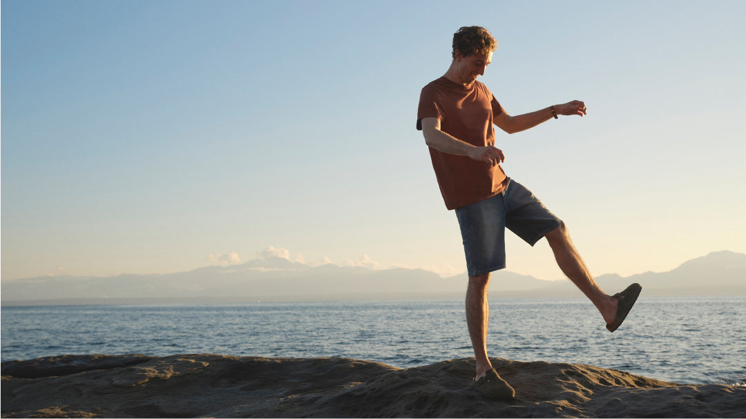 A man wearing a brown t-shirt and jean shorts stands on a rocky shoreline, with mountains and the ocean behind them. 